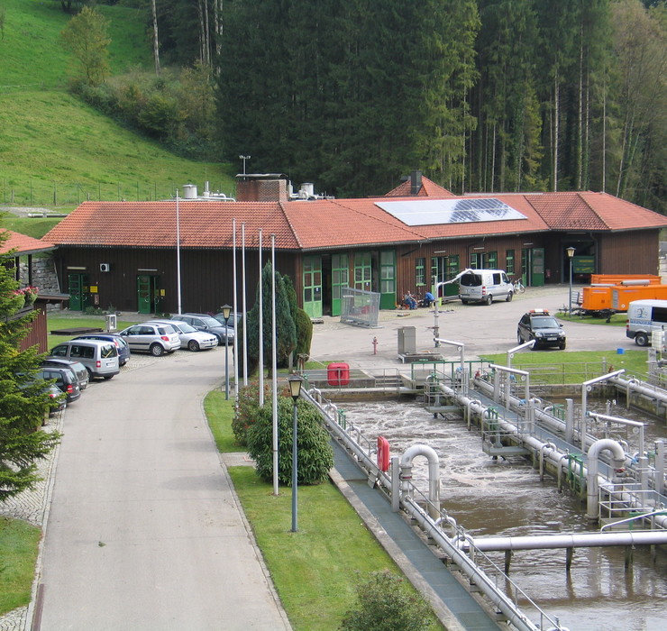AUV-Chiemsee Kläranlage beim AUV-Chiemsee, im Vordergrund ein klärbecken, hinten ein Gebäude mit Solarpanelen, davor stehen Autos und ein Mitarbeiter sitz auf der Bank vor dem Gebäude